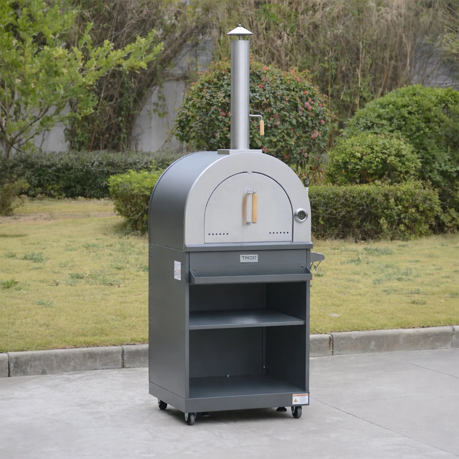 Outdoor stainless steel pizza oven with chimney and shelves on wheels in a garden setting