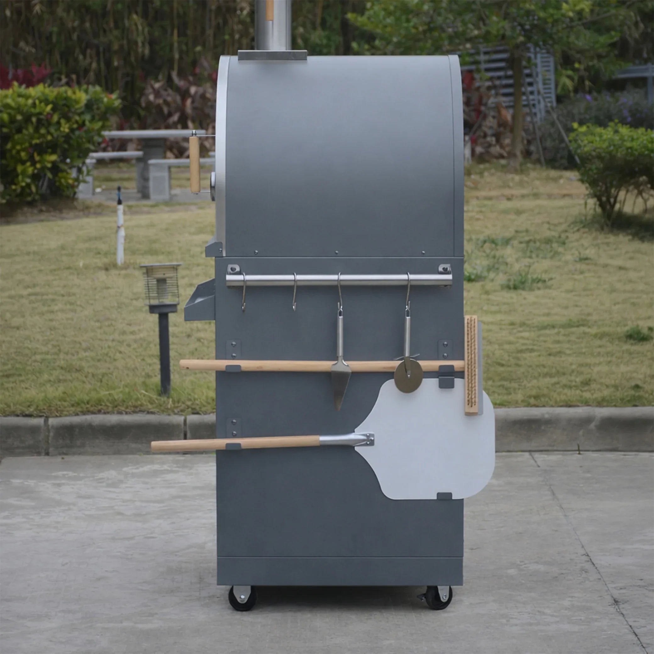Outdoor wood-fired pizza oven with tools on wheels in a garden setting