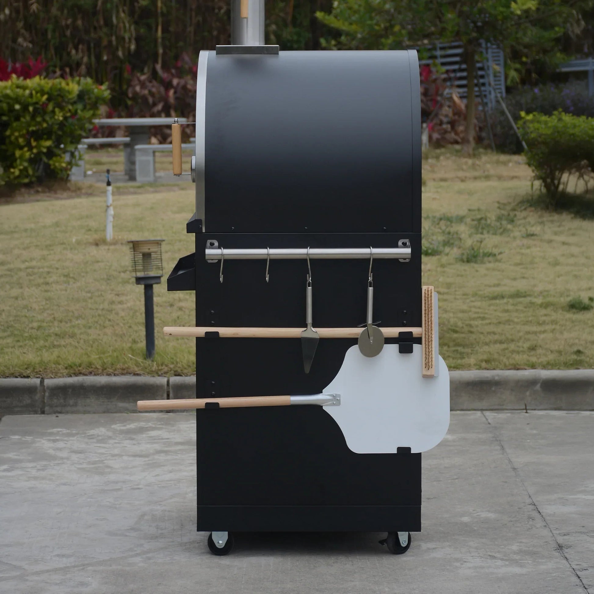 Outdoor black pizza oven with wood handles, pizza peel, and utensils on wheels in a garden setting