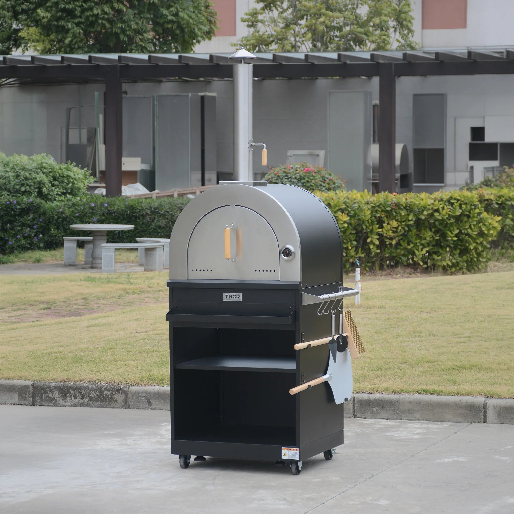 Outdoor stainless steel wood-fired pizza oven with chimney and cooking tools on wheels