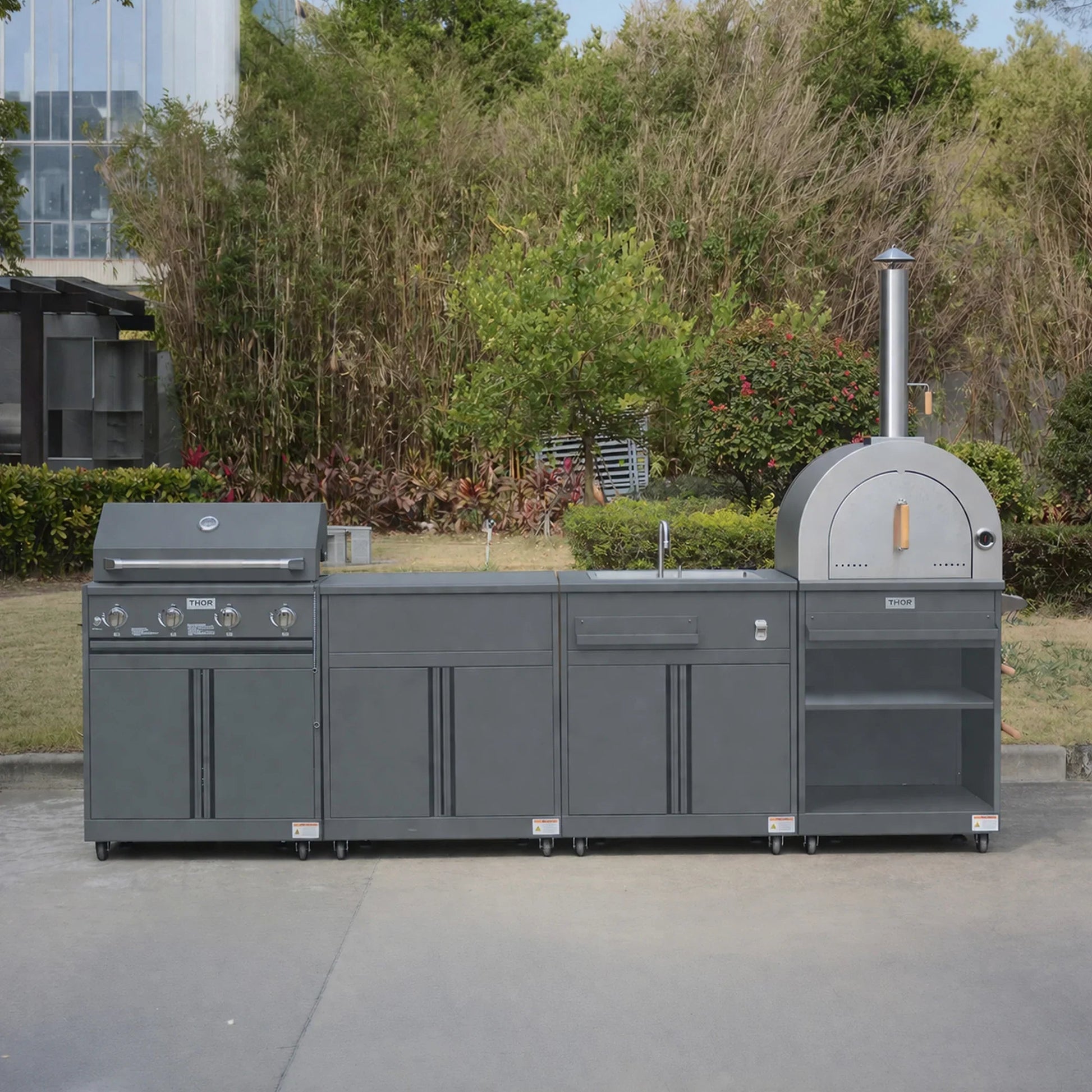 Modern outdoor kitchen set with grill, pizza oven, sink and storage, in a garden setting.